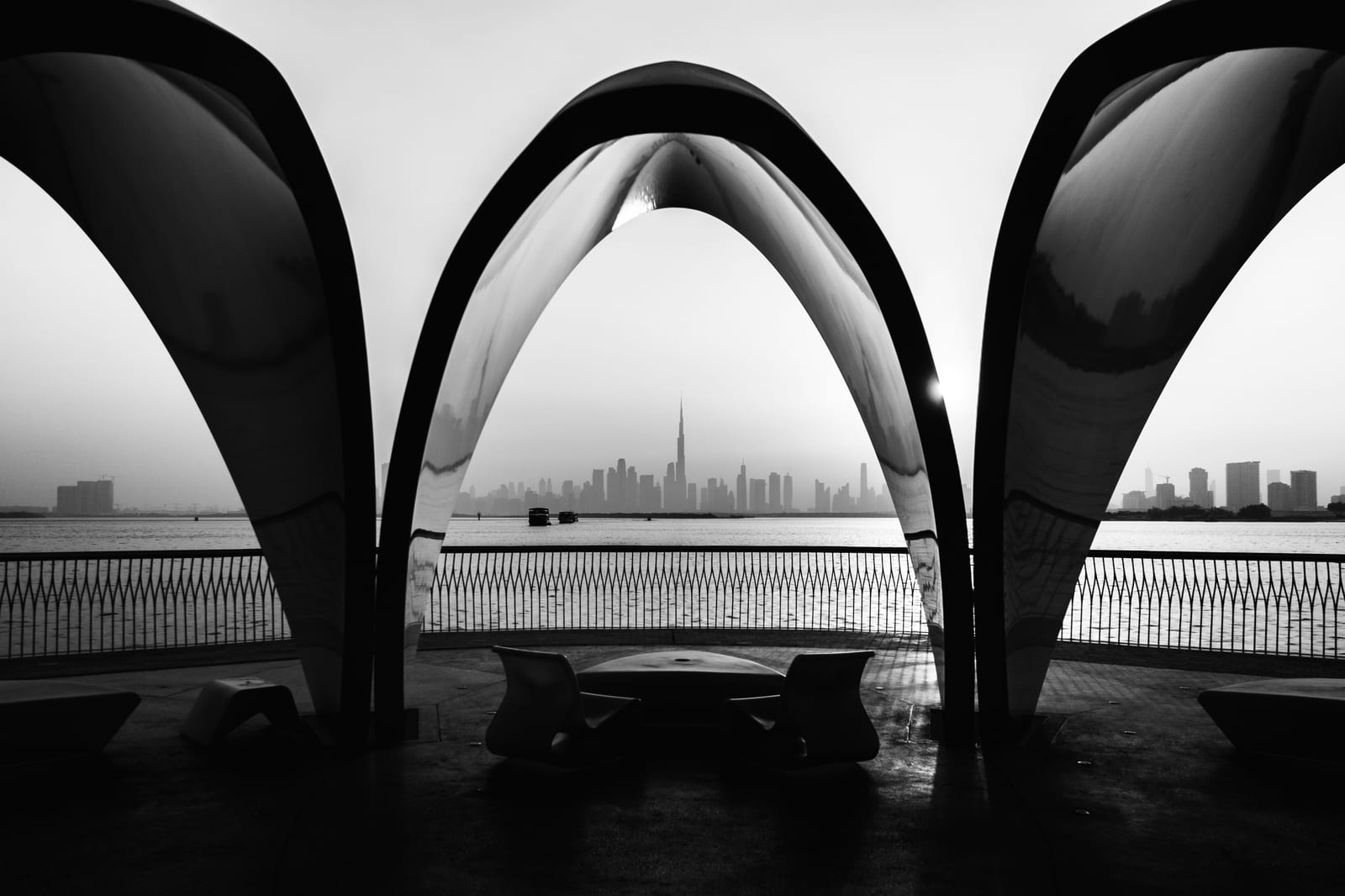 A distant city skyline across the water, framed by the arches of a waterfront shelter